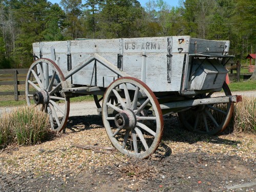 An old wooden wagon with large wheels, labeled "U.S. Army," sits on gravel surrounded by greenery.