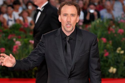 A man in a black suit gestures while walking on a red carpet, surrounded by a crowd and floral decorations.