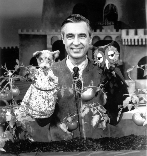 A man smiles with two puppets in front of tomato plants, set in a whimsical garden backdrop.