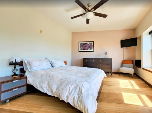 Bright bedroom with a queen bed, dresser, TV, and large window, featuring bamboo flooring and soft pastel walls.