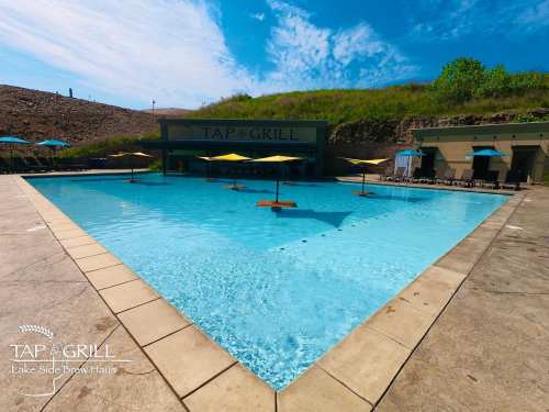 A serene pool area with umbrellas, surrounded by greenery and a building labeled "Tap & Grill." Clear blue sky above.