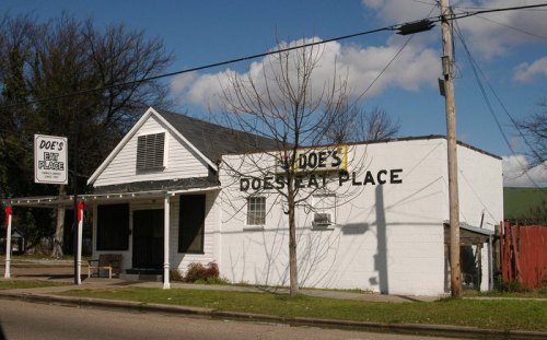 A small, white building with a sign reading "Doe's Eat Place," surrounded by trees and a clear blue sky.