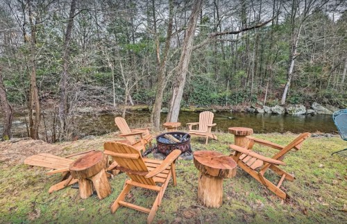 A circle of wooden chairs around a fire pit by a serene stream, surrounded by trees and greenery.