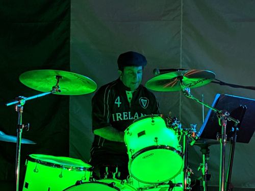 A drummer in a green-lit setting, wearing an Ireland jersey, plays on a drum set with a focused expression.