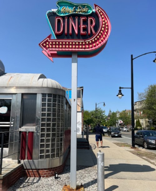 Neon sign for "West Side Diner" with an arrow, set against a clear blue sky and a street scene.