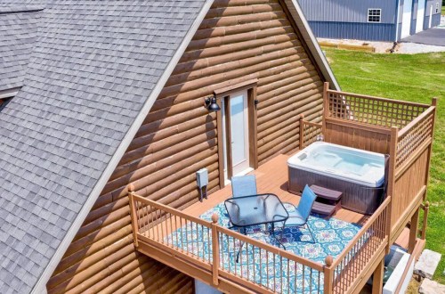 Aerial view of a wooden cabin with a deck featuring a hot tub and outdoor seating on a sunny day.