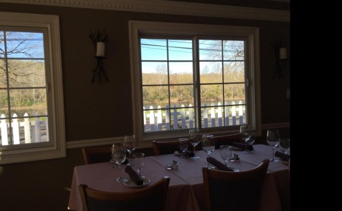 A dining table set with glasses and napkins, with large windows showing a view of trees and a white fence outside.