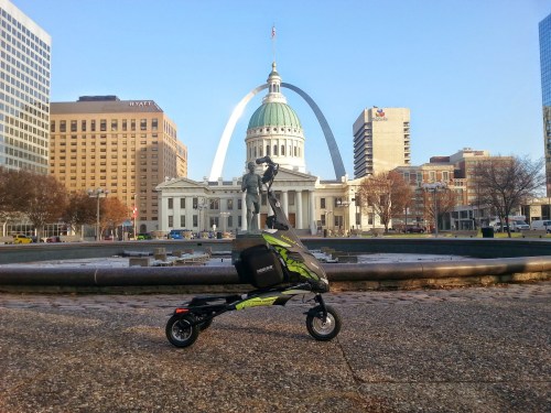 A scooter parked in front of a historic building and the Gateway Arch in a cityscape.