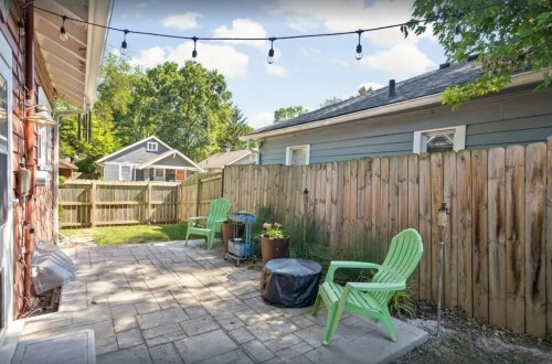 A cozy backyard patio with green chairs, a fire pit, and string lights, surrounded by a wooden fence and trees.