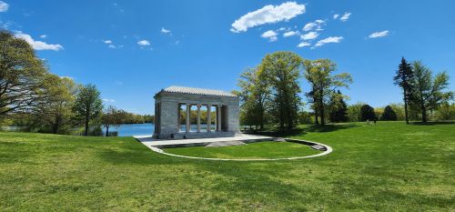 A stone monument with columns surrounded by green grass and trees, near a calm lake under a blue sky with clouds.