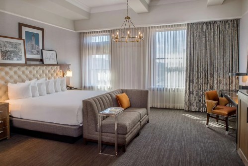 A stylish hotel room featuring a king bed, seating area, large windows, and modern decor.