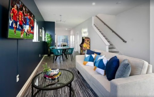 A modern living room with a sofa, coffee table, and a TV showing a soccer game, featuring a dining area and staircase.