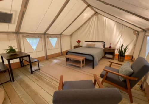 Cozy glamping tent interior with a bed, seating area, and wooden furniture, surrounded by soft lighting.