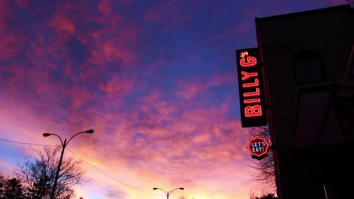 A vibrant sunset sky with colorful clouds, featuring a restaurant sign that reads "BILLY G's" and "LET'S EAT."