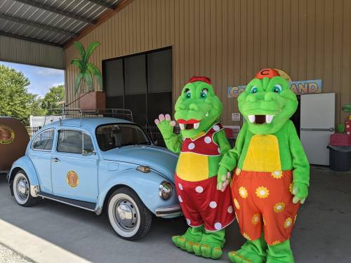 Two green alligator mascots in colorful outfits stand beside a blue vintage car in a playful setting.