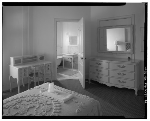 A vintage bedroom with a white bedspread, dresser, and a desk, featuring an open door leading to another room.