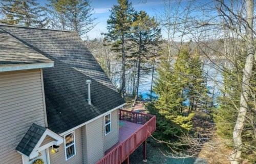 Aerial view of a house with a deck, surrounded by trees near a calm lake on a clear day.