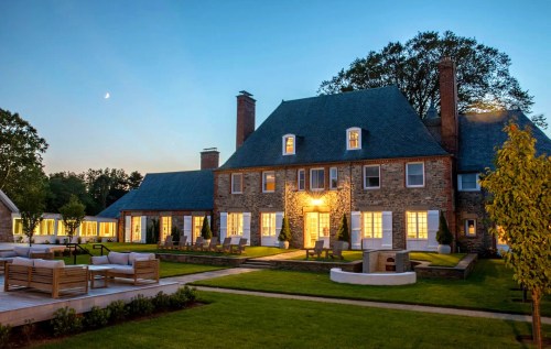 A large stone mansion at dusk, surrounded by green lawns and outdoor seating, with warm lights glowing from the windows.