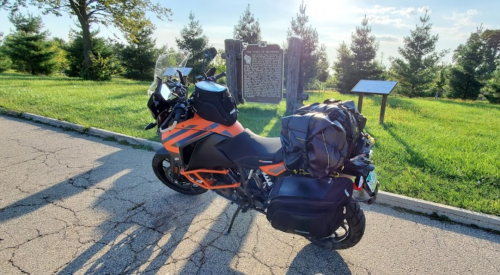 A motorcycle parked on a cracked road with trees and historical markers in the background under a clear sky.