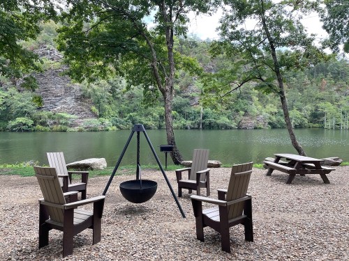 A serene lakeside scene with four wooden chairs around a fire pit, surrounded by trees and rocky cliffs.