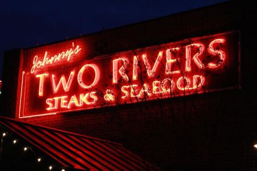 Neon sign for "Johnny's Two Rivers Steaks & Seafood" glowing against a dark evening sky.