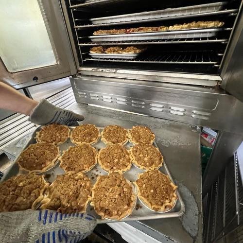 A person wearing oven mitts pulls a tray of apple pies from a commercial oven, with more pies visible inside.