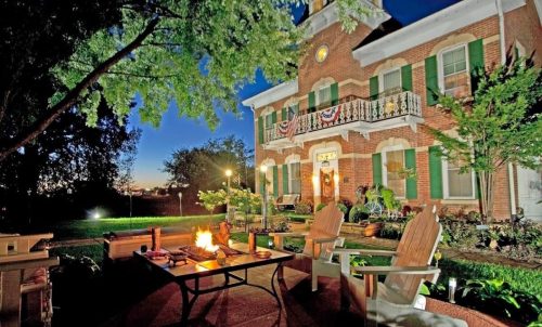 A cozy outdoor seating area with a fire pit in front of a charming brick house, illuminated at dusk.