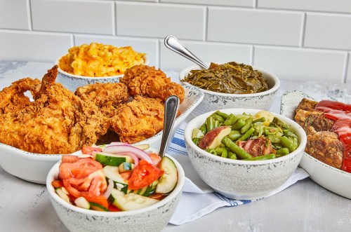 A spread of Southern comfort food including fried chicken, mac and cheese, collard greens, and mixed vegetables in bowls.