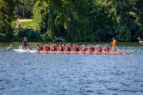 A dragon boat with rowers in life vests glides across a calm river, surrounded by lush greenery.