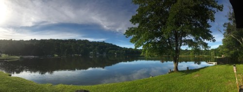 A serene lake surrounded by lush greenery and trees, reflecting the sky and hills in calm waters.