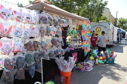 A vibrant market stall displays colorful masks and toys, with children browsing and a trailer in the background.