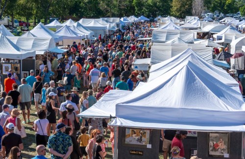 A bustling outdoor market with numerous white tents and a large crowd of people browsing art and crafts.