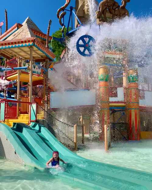 A child splashes in a water park slide as water cascades down from above, surrounded by colorful attractions.