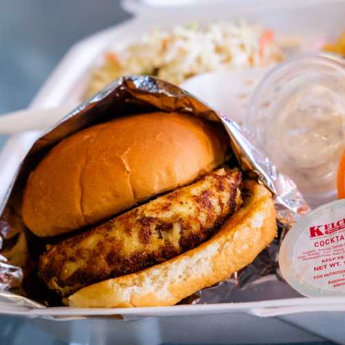 A close-up of a fried chicken sandwich in a bun, served with coleslaw and a small container of cocktail sauce.