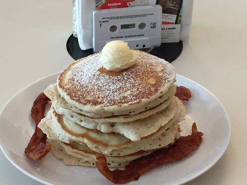 A stack of fluffy pancakes topped with butter and powdered sugar, served with crispy bacon on the side.