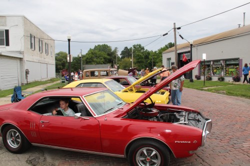A red Pontiac car with its hood open, parked on a brick street alongside other classic cars and people enjoying the event.