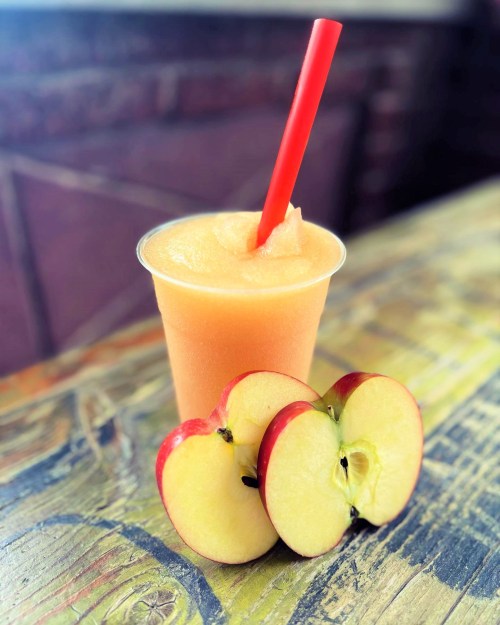 A frozen drink in a cup with a red straw, accompanied by two sliced red apples on a wooden table.
