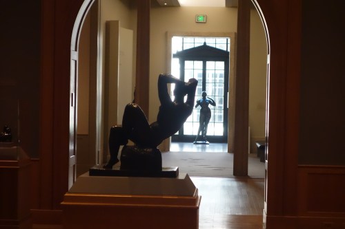 A sculpture of a seated figure in a gallery, with another figure visible in the background through an archway.