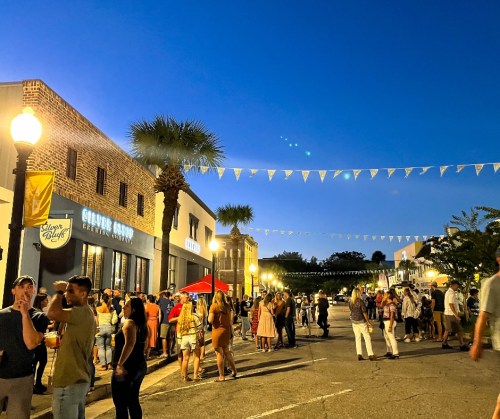 A lively street scene at dusk with people socializing, string lights, and palm trees in a festive atmosphere.