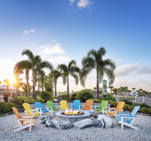 Colorful Adirondack chairs surround a fire pit, set against a backdrop of palm trees and a clear blue sky.