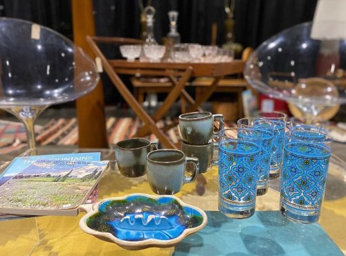 A table with colorful glasses, ceramic dishes, and a magazine, set in a cozy, vintage-style environment.