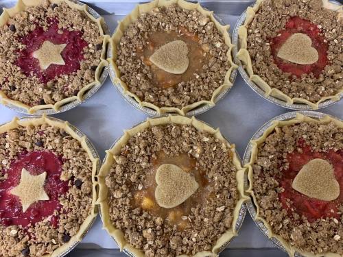 Six pies with crumbly toppings and decorative shapes, arranged on a baking tray. Flavors include cherry and peach.