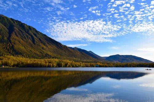 A serene lake reflects mountains and a blue sky with scattered clouds, surrounded by autumn-colored trees.