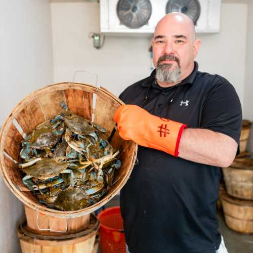 A man in a black shirt holds a basket of blue crabs, with wooden barrels in the background.