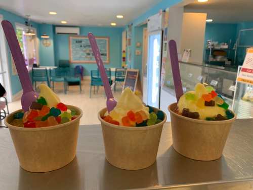 Three cups of soft serve ice cream topped with colorful gummy candies, set in a bright, inviting ice cream shop.