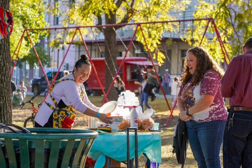 A woman serves food at a table in a park, with swings and people enjoying the day in the background.