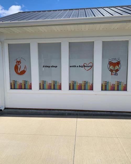 Storefront with large windows displaying colorful book graphics and text: "A tiny shop with a big heart" and "Swampy Fox Kids."