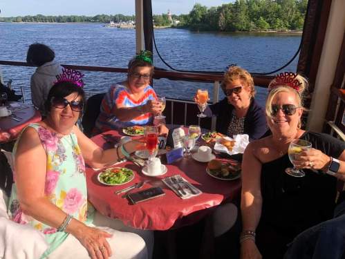 Four women celebrate a birthday at a table by the water, enjoying drinks and food, wearing festive party hats.