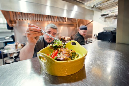 A chef carefully garnishes a colorful dish in a busy kitchen, with another chef working in the background.