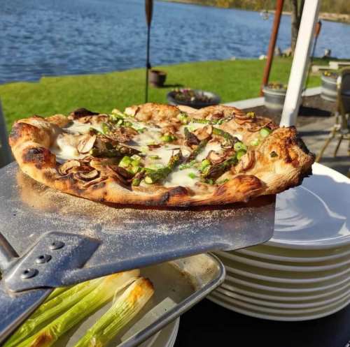 A freshly baked pizza with mushrooms and green onions, held above a table by a scenic lakeside.